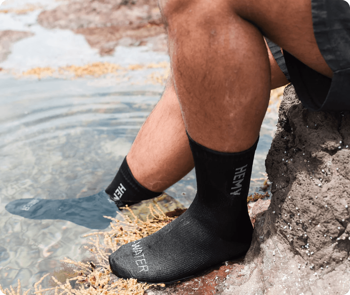 Close up of a man sitting on a rock wearing Hemy socks, with one foot in water